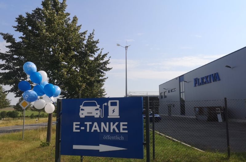 Im Vordergrund steht ein Schild mit der Aufschrift "E-Tanke öffentlich" im Hintergrund steht ein Gebäude mit der Aufschrift "FLEXIVA"